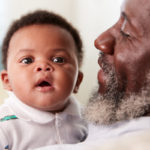 Proud Grandfather Cuddling Baby Grandson In Nursery At Home
